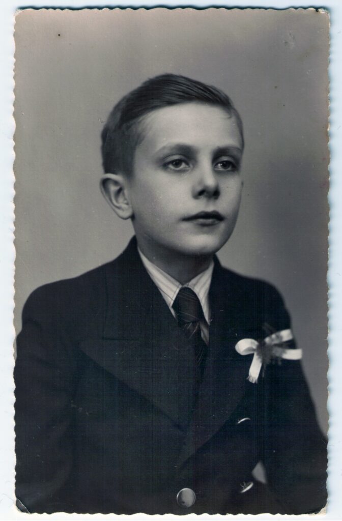 Czarno-biała fotografia portretowa przedstawiająca kilkuletniego chłopca ubranego w marynarkę z krawatem. Chłopiec patrzy lekko w bok, ma krótkie, starannie uczesane włosy i poważny wyraz twarzy. Zdjęcie ma ozdobnie ząbkowane brzegi.